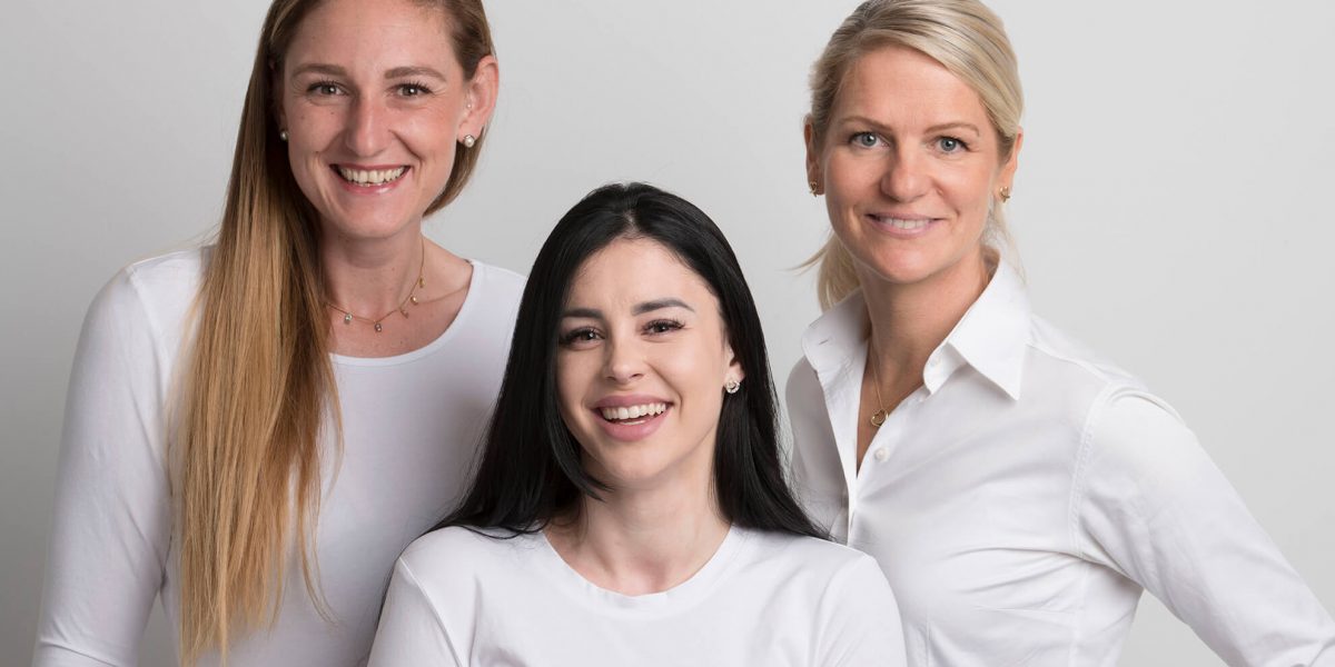 Teamfoto mit drei Mitarbeiterinnen in weißem Oberteil.